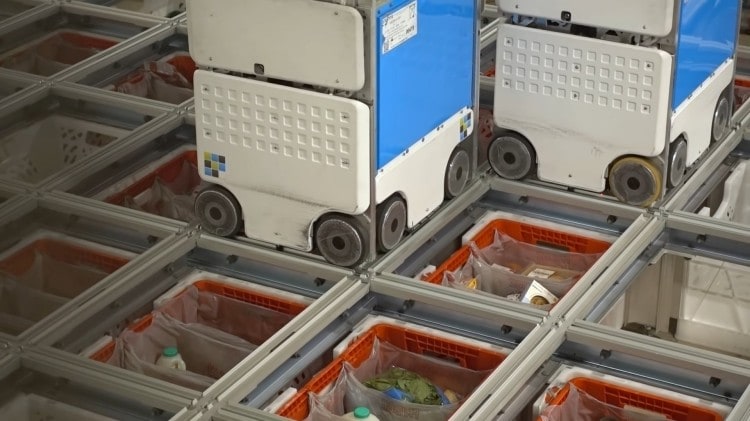 오카도 풀필먼트 창고의 패키징 로봇들이 배송 바구니에 성품을 투하하고 있다